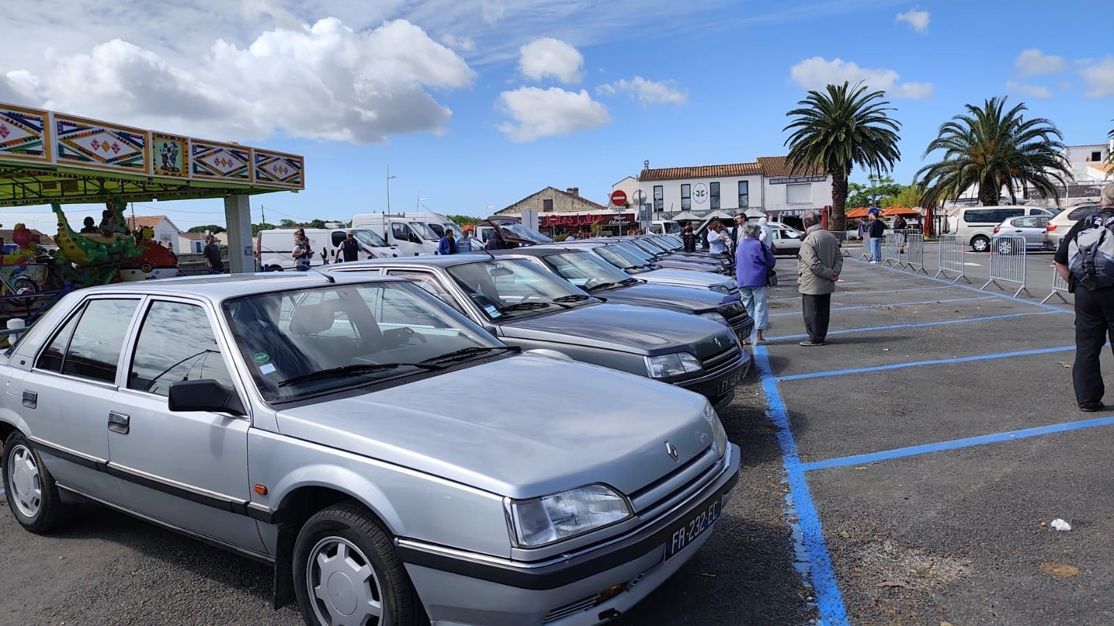 A grey Renault 25 in a car park, in front of a market and palm trees